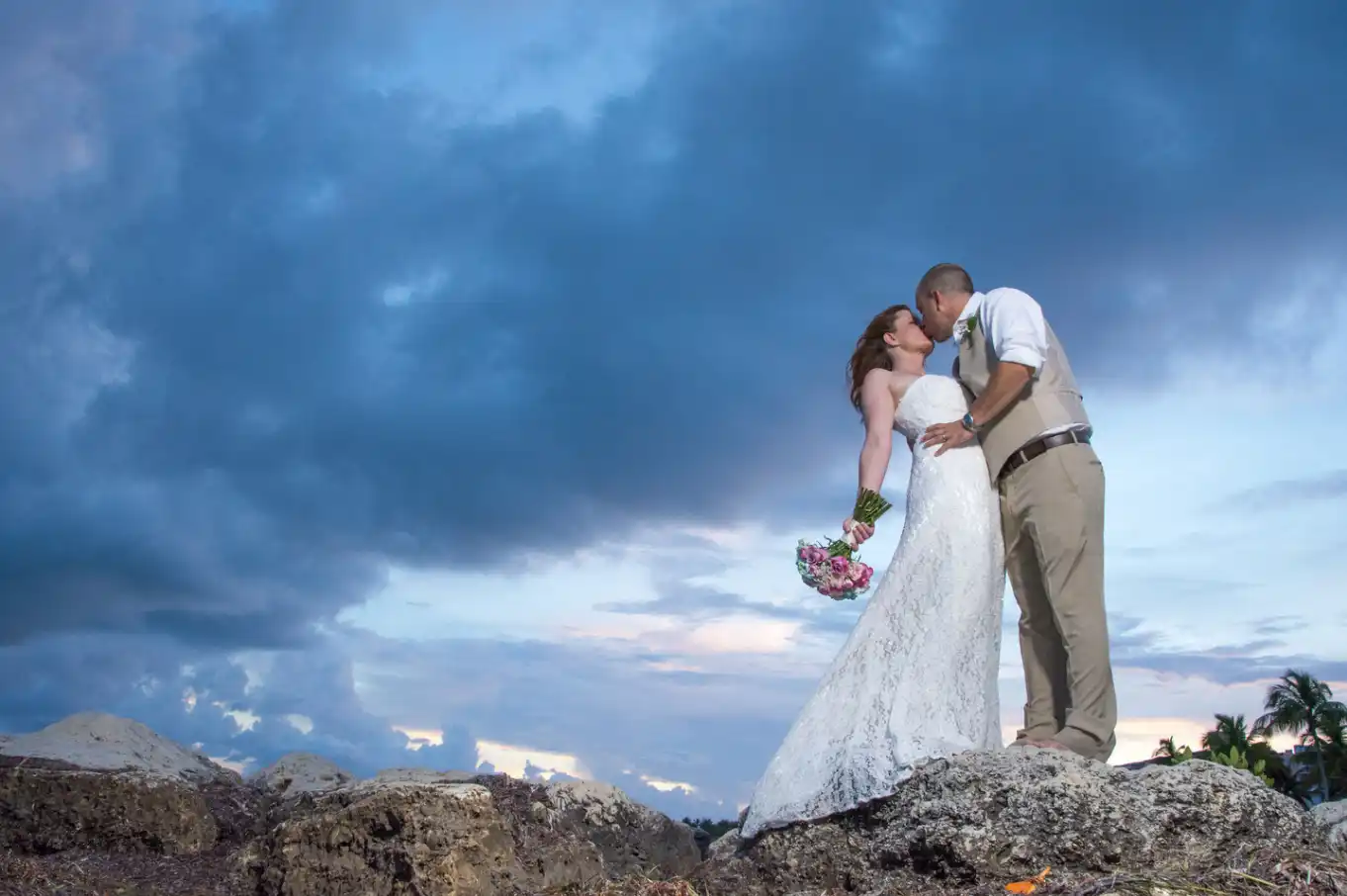 A key west bride and groom kissing on rocks in front of a cloudy sky.