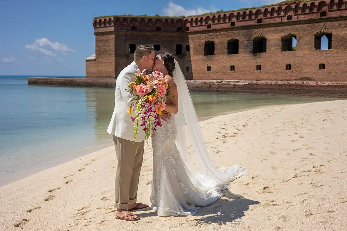 As a key west wedding photographer, I specialize in capturing magical moments like a bride and groom kiss on the beach in front of an old fort. With my expertise, I can provide you with stunning photographs