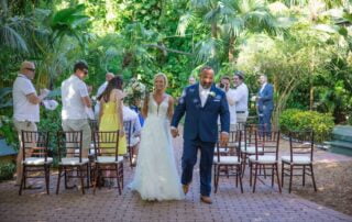 A bride and groom adorned in matching floral boutonnieres and a delicate white lace wedding dress, joyfully stroll down the flower-lined aisle of a picturesque tropical garden.