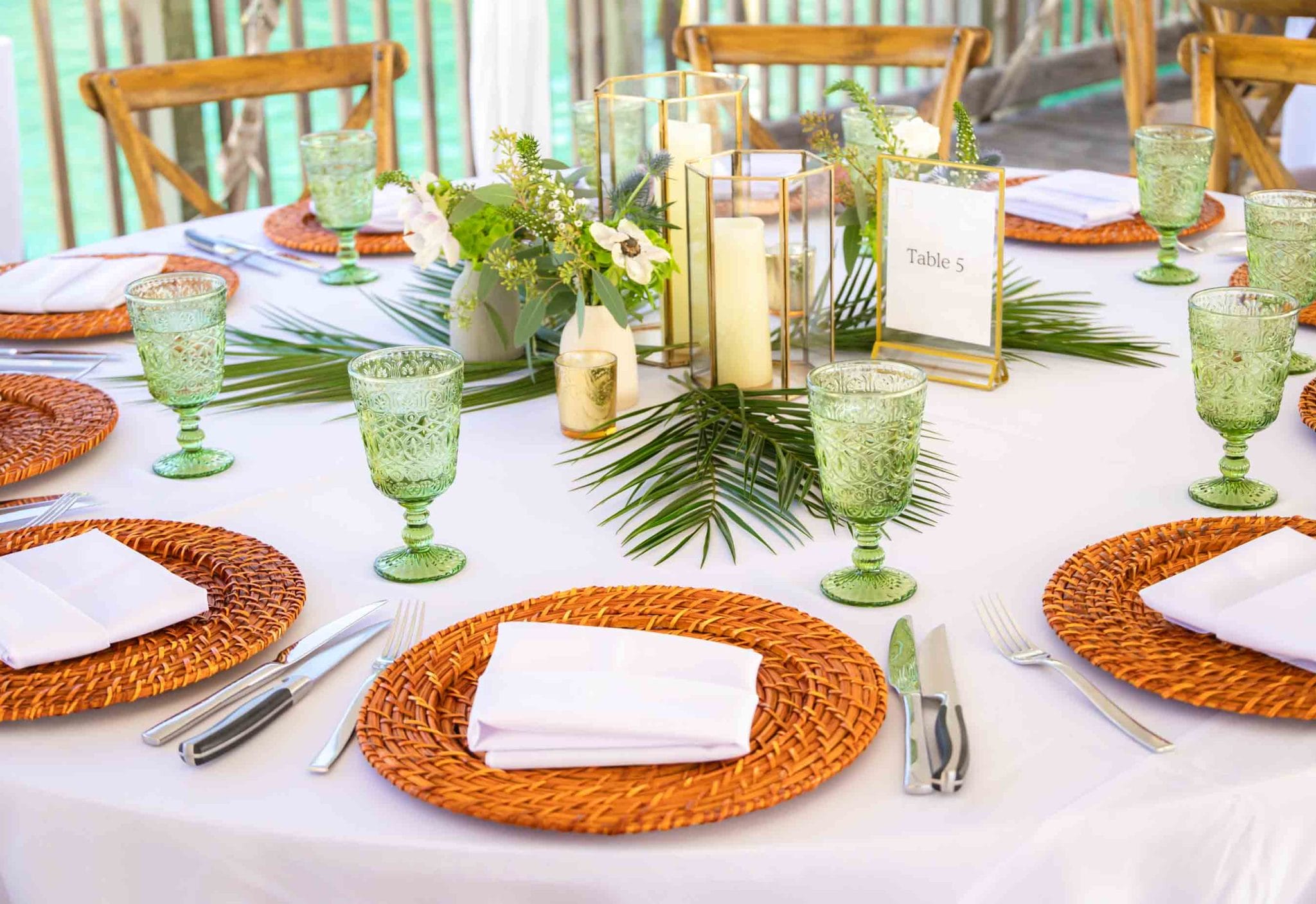The image shows a table setting from a top-down perspective, part of a larger event setup. There's a focus on natural and earthy tones. A white tablecloth serves as the base, with a centerpiece composed of green tropical leaves, white candles of varying heights, and clear glassware. Each place setting has a woven rattan charger, white dinner plate, green glass goblet, and silverware. The style suggests a tropical, elegant theme, likely for an event such as a wedding or upscale dinner. The setting exudes a relaxed yet sophisticated ambiance, potentially fitting for a destination event in a warm, beachside location.