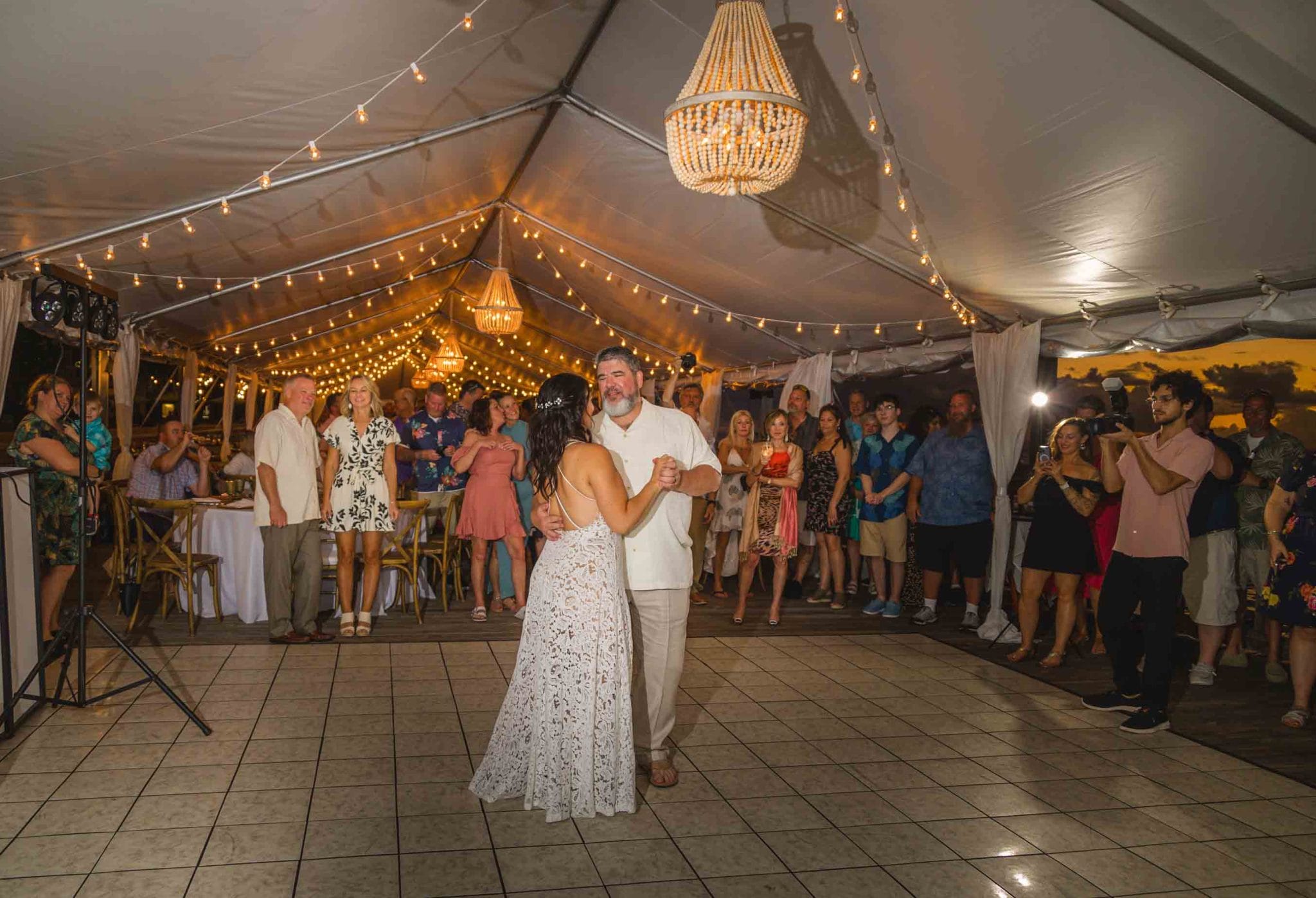 This image captures a joyous wedding reception at the Ocean Key Resort and Spa in Key West. The bride and groom are in the center of the dance floor, sharing a tender moment, surrounded by guests. The venue is adorned with string lights creating a warm, inviting glow that complements the dusky evening sky visible through the open sides of the tent. The scene is lively yet intimate, with the elegant attire of the guests adding to the festive atmosphere. The oceanfront setting, combined with the sophisticated decor, offers a glimpse into a memorable celebration at this destination wedding.