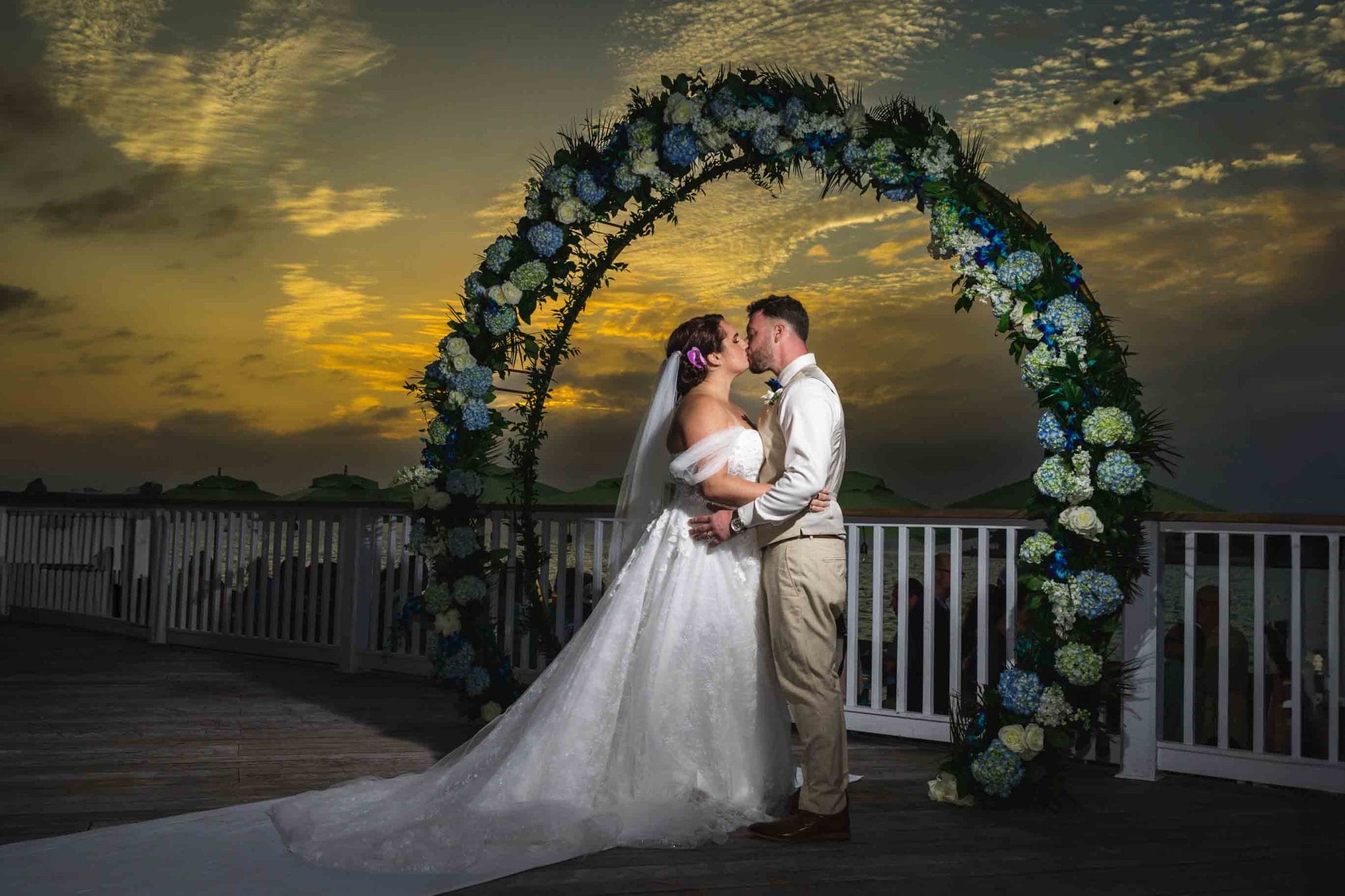 A bride and groom kissing under a floral arch, with a dramatic Key West sunset sky in the background.