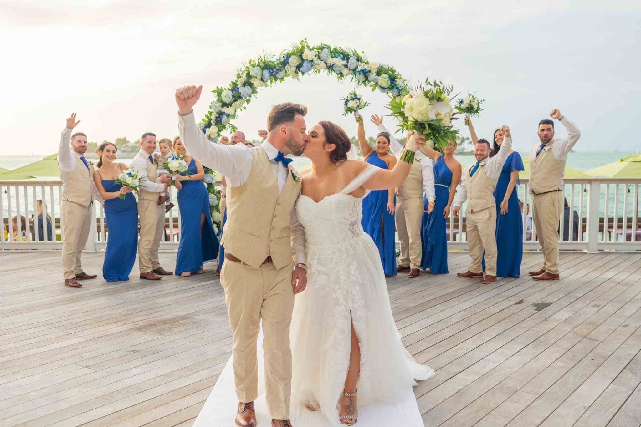 A bride and groom kiss on a wooden pier with their wedding party, wearing blue and beige, cheering behind them under a floral arch.