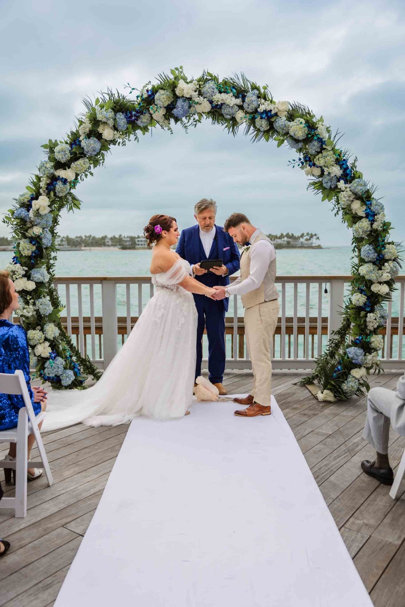 A bride and groom exchange rings at an outdoor wedding ceremony by the sea, under a floral archway, with an officiant and guests seated nearby.