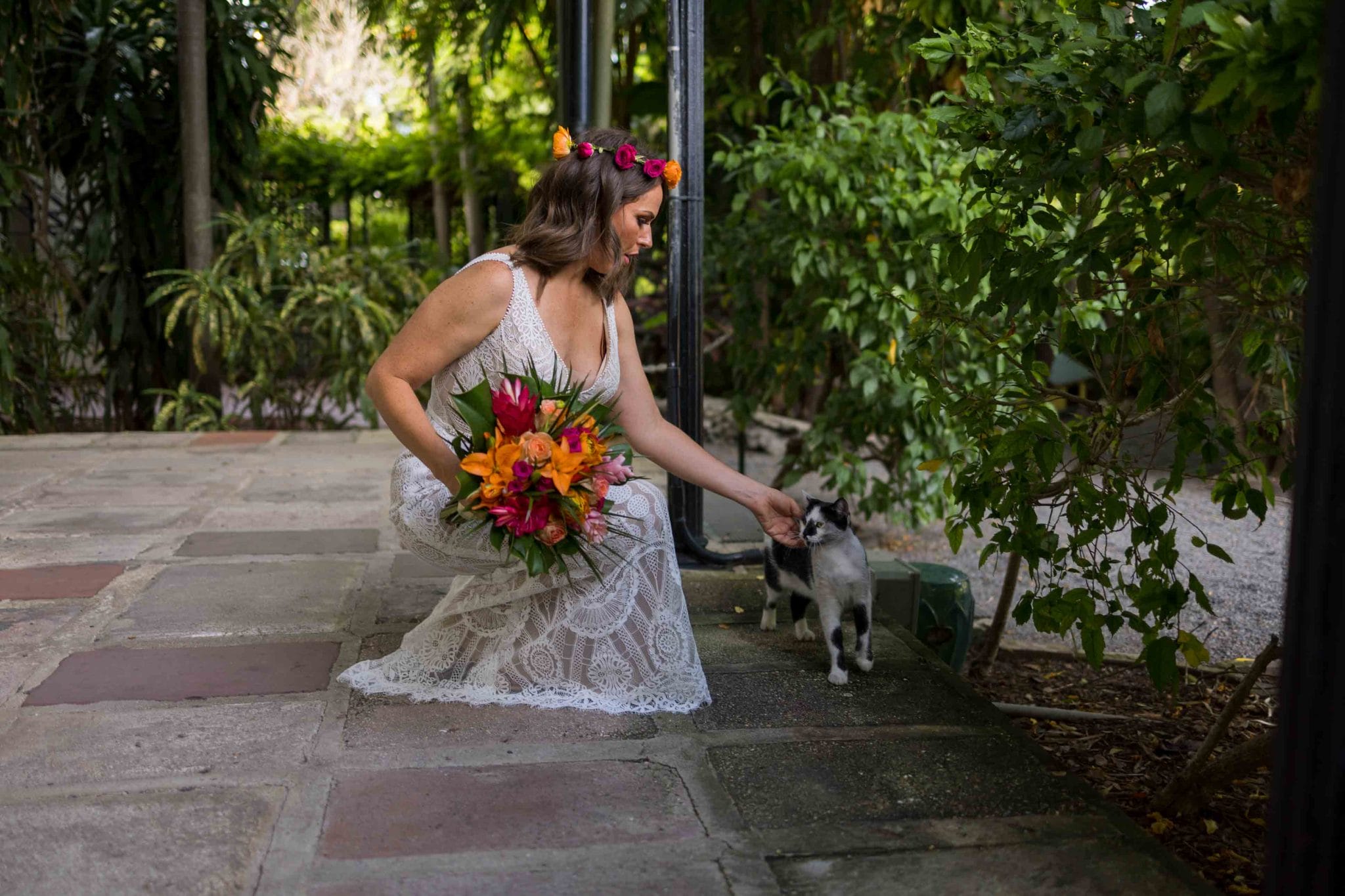 Hemingway Home Wedding A woman in a white lace dress and floral headband pets a cat on a stone path surrounded by lush greenery, holding a bouquet of bright flowers.