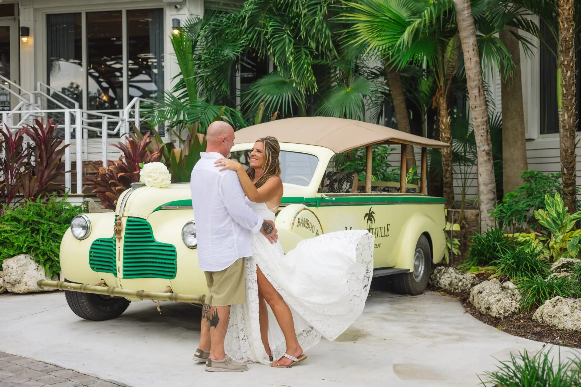 A bride and groom embrace in front of a vintage cream-and-green bus with tropical palm trees in the background.