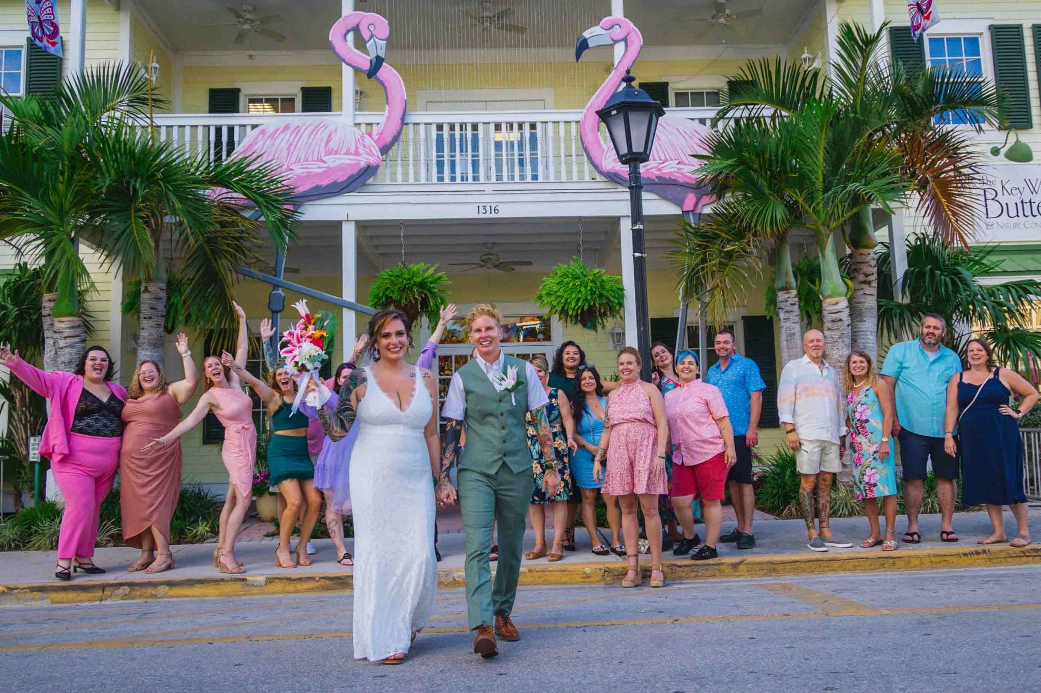 A joyful wedding scene with a couple walking hand in hand down a street, flanked by friends and large flamingo decorations in front of a white house.