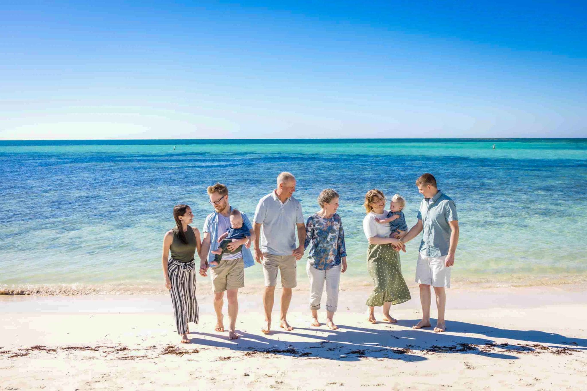 A group of seven people, including two babies, walking and chatting on a sandy beach with blue ocean and sky in the background.