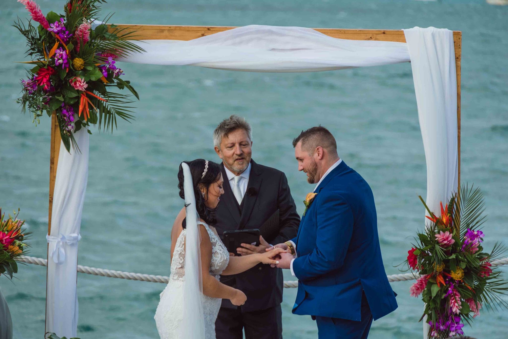 Hyatt Wedding by Michael Vernon Officiant A couple exchanges rings in an outdoor wedding ceremony by the sea, under a wooden arch with white fabric and colorful flowers, while an officiant stands behind them.