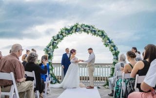 Couple exchanging vows under a floral arch at an outdoor wedding ceremony by the sea, attended by guests seated on white chairs.