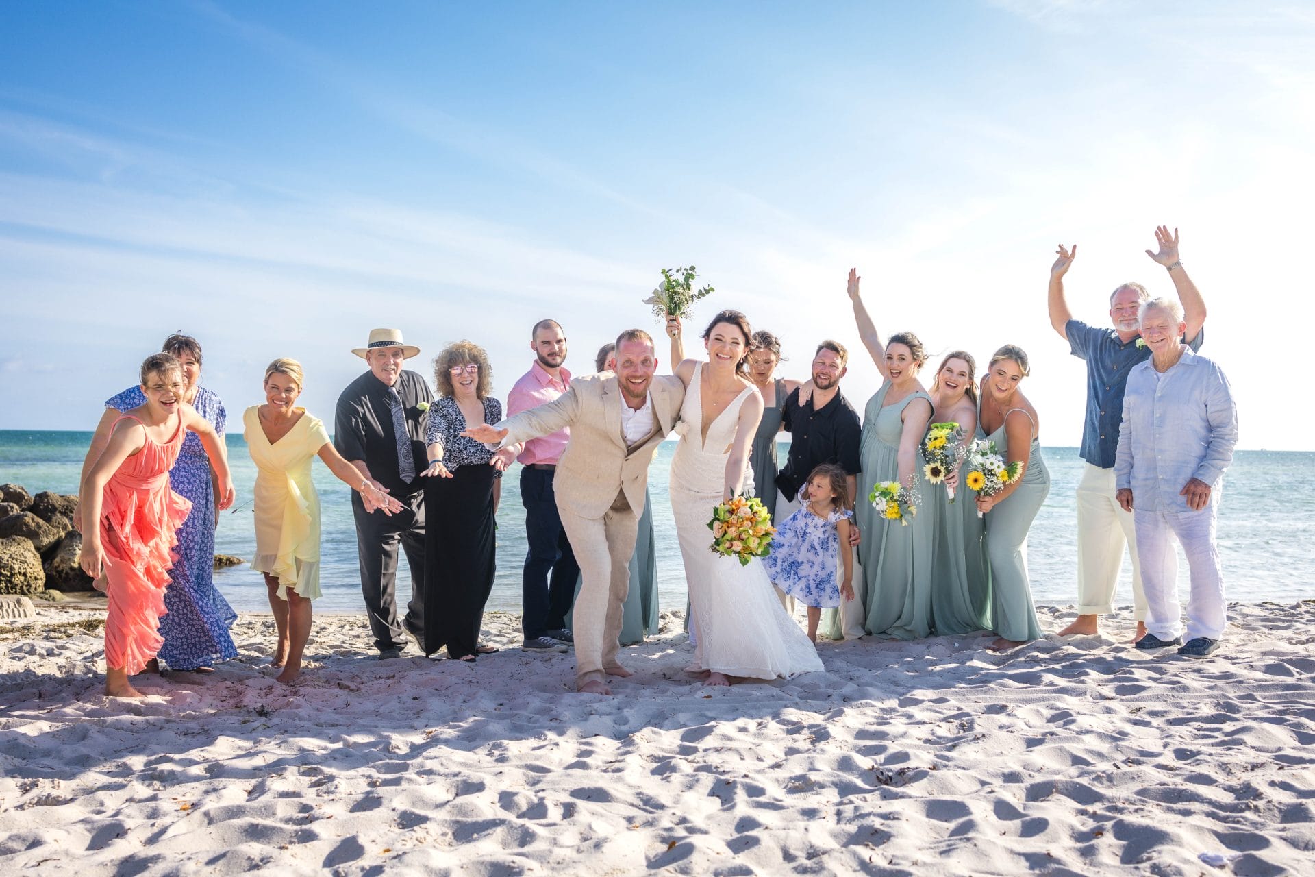 A wedding party poses joyfully on a sandy beach with a blue sky and ocean in the background. The bride and groom stand in the center, surrounded by friends and family.