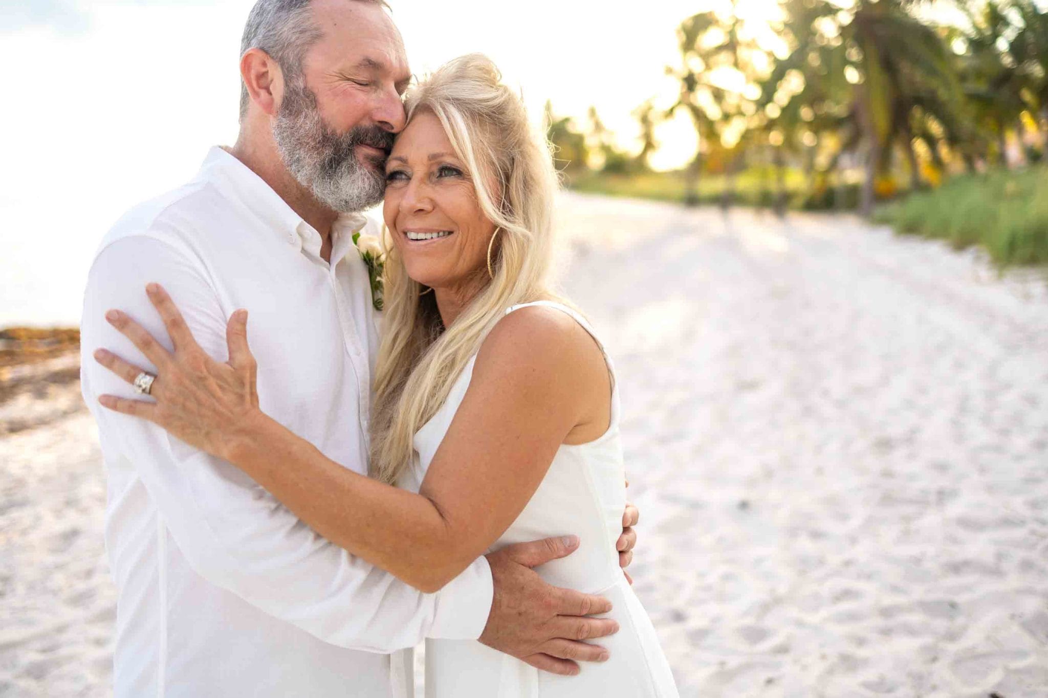 A couple embraces on a sandy beach lined with palm trees under a clear sky.