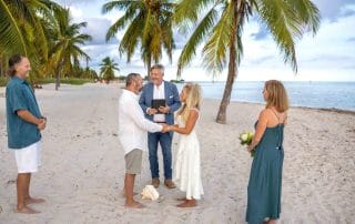 A couple holds hands during a beach wedding ceremony with two guests and a person officiating under palm trees.