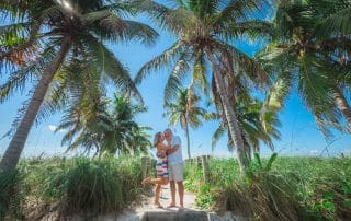 A couple embraces under palm trees on a sunny beach, surrounded by lush greenery.