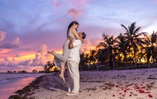 A couple on a beach at sunset, with the man lifting the woman. Palm trees and rose petals are visible in the background.