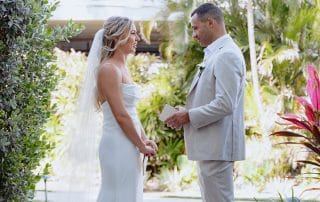 Bride and groom standing outside in a garden, exchanging vows. Both wear white; the groom is in a light suit and the bride in a dress with a veil. Tropical plants surround them.