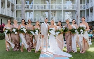 A wedding party poses outdoors in front of a building. The groom is flanked by nine bridesmaids in matching dresses, holding bouquets.