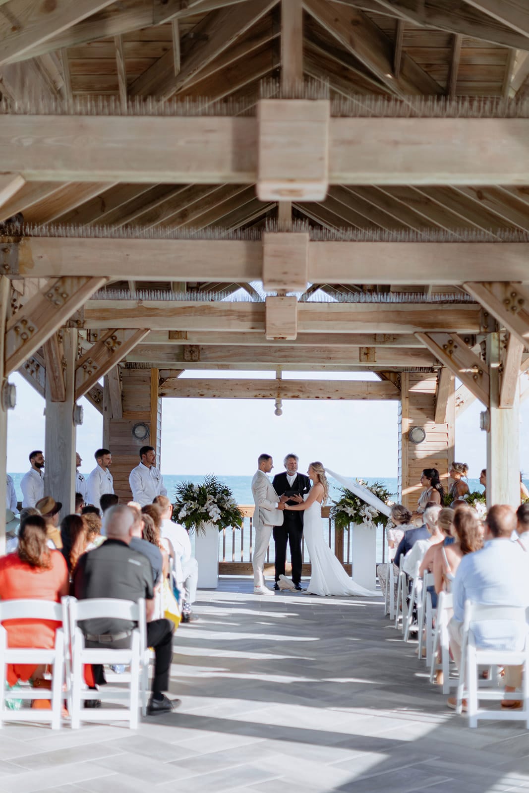 Courtney & Spencer A couple is getting married under a wooden pavilion by the ocean, with guests seated on white chairs observing the ceremony. performed with Michael Vernon