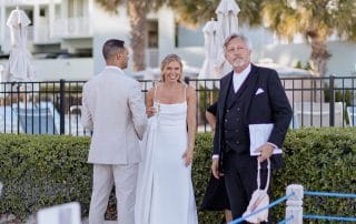 Three people in formal attire stand outdoors near a bush fence. The woman in a white dress holds a glass, smiling. The man beside her wears a light suit, and the man on the right wears a dark suit and holds papers.