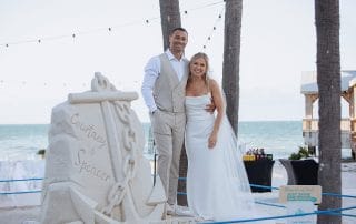 A couple in wedding attire stands by an anchor-shaped sand sculpture with their names. The beach and ocean are in the background.
