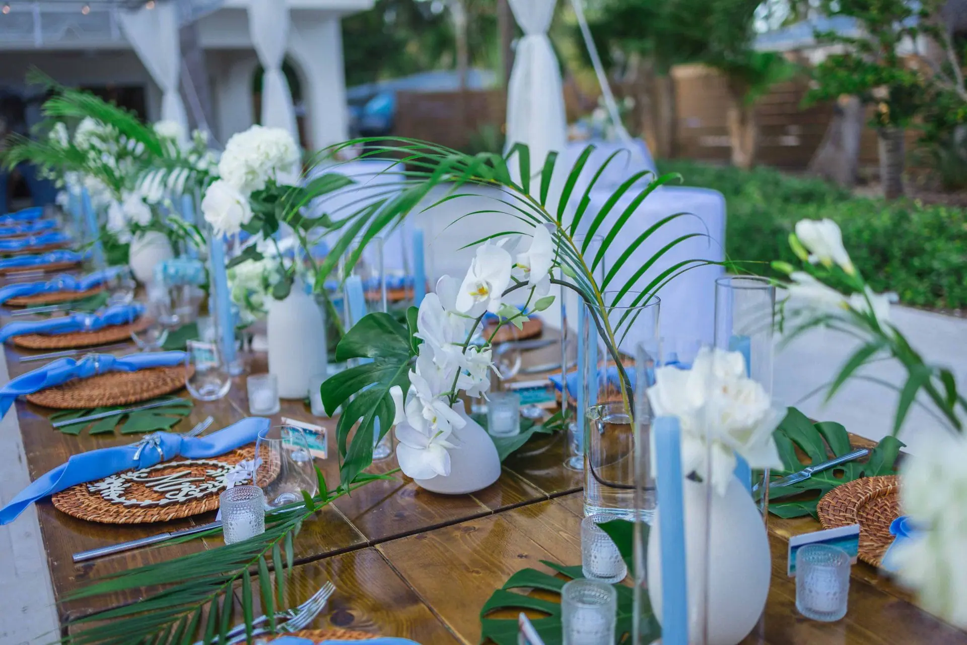 A long wooden table is set for an outdoor event, decorated with white flowers, green leaves, and blue napkins.