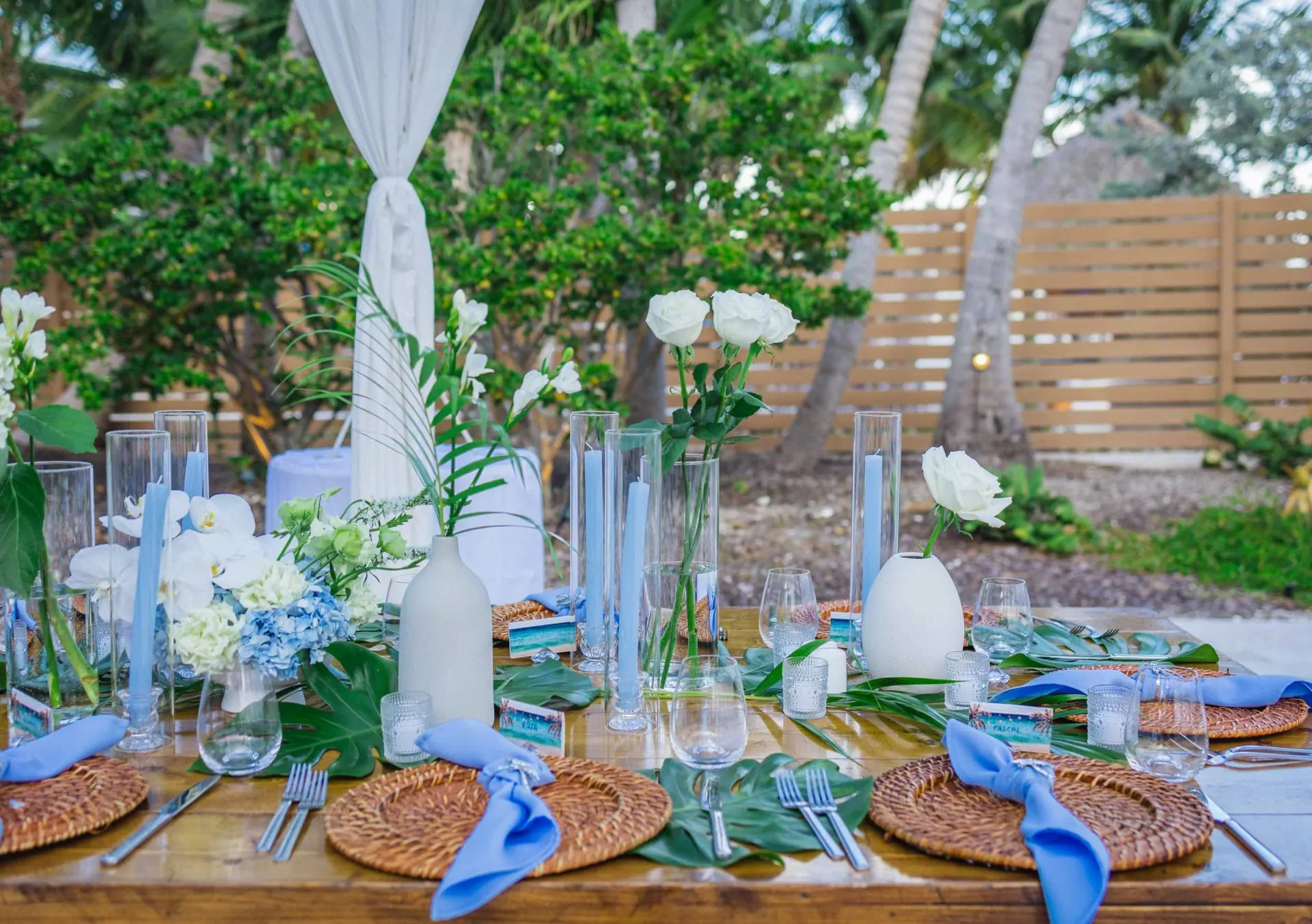 Outdoor table setting with woven placemats, blue napkins, clear glasses, and white floral centerpieces. Greenery in the background under a white canopy.