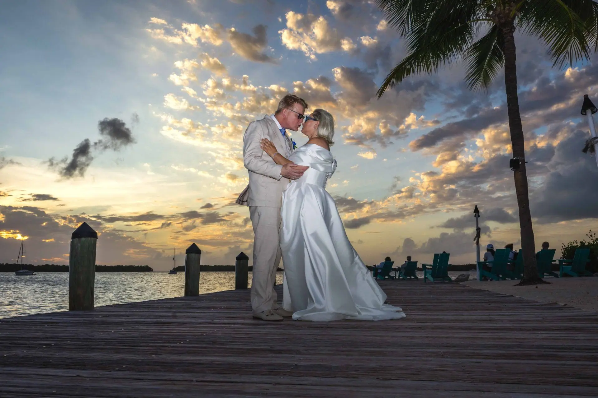 A couple in formal attire embraces on a wooden dock by the water during sunset, with clouds and a palm tree in the background.