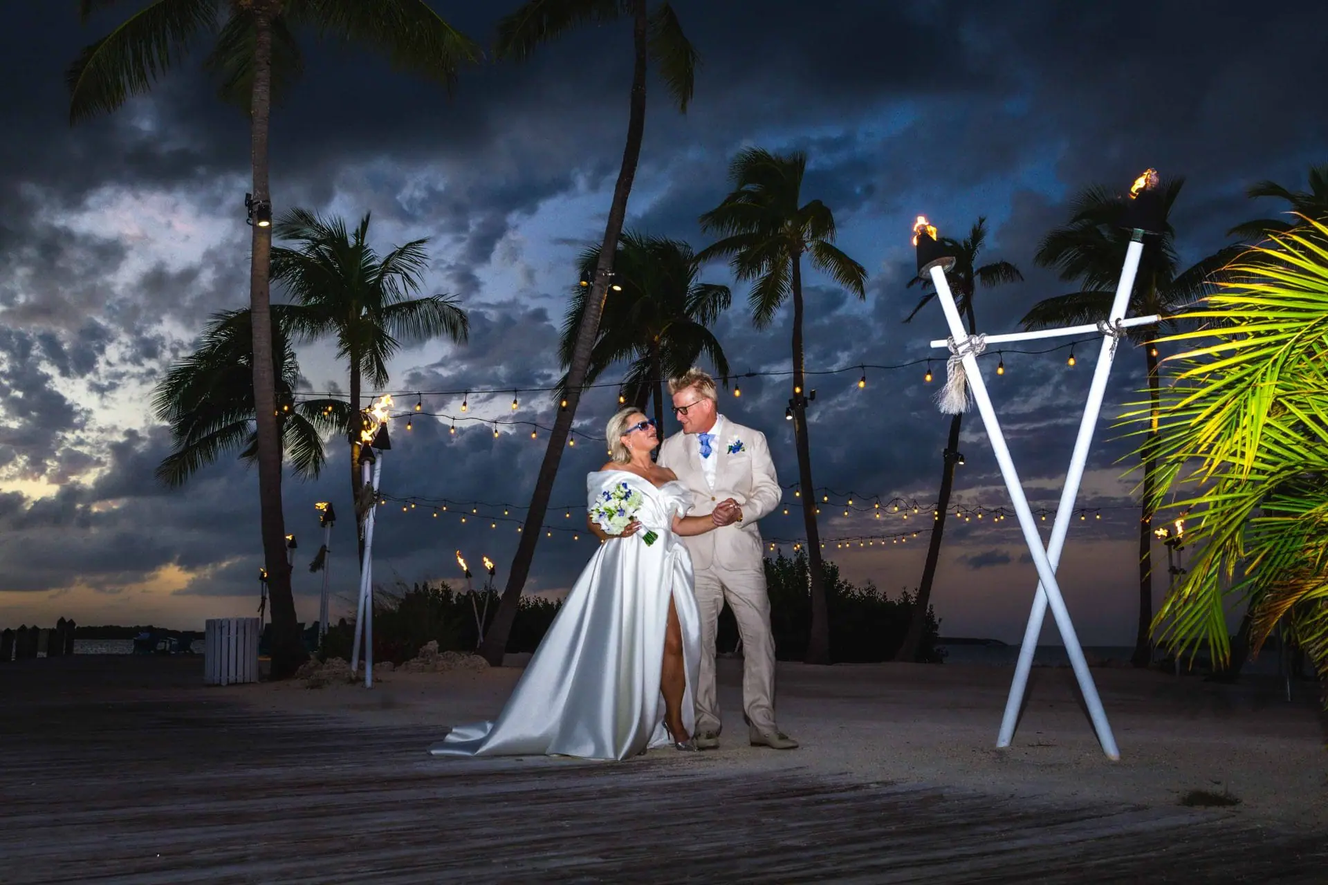 A bride and groom stand under palm trees and string lights on a wooden deck at dusk, with tiki torches and a cloudy sky in the background.
