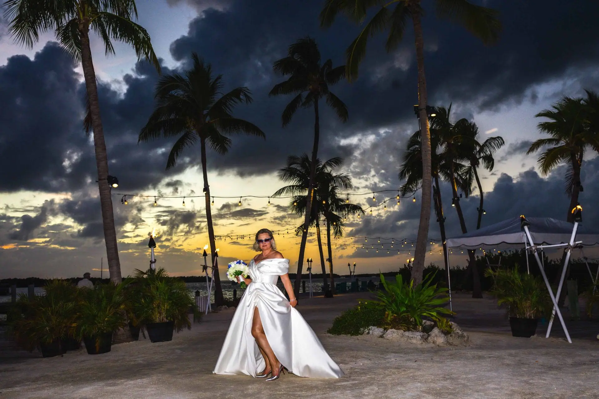 Bride in a white dress poses in an outdoor setting with palm trees and a sunset sky.