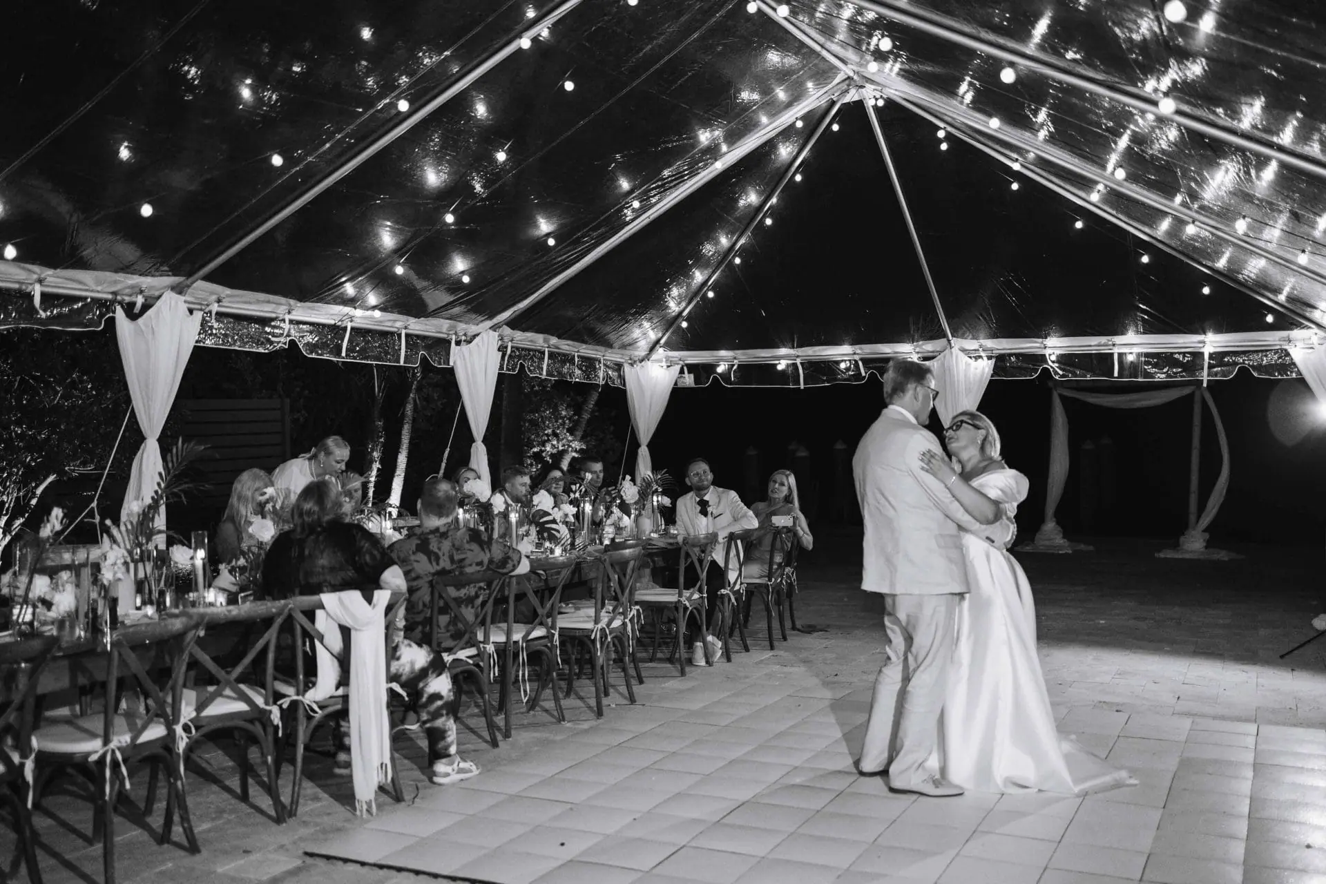 A couple shares a first dance under a string-lit tent at a wedding reception, while guests seated at tables watch.