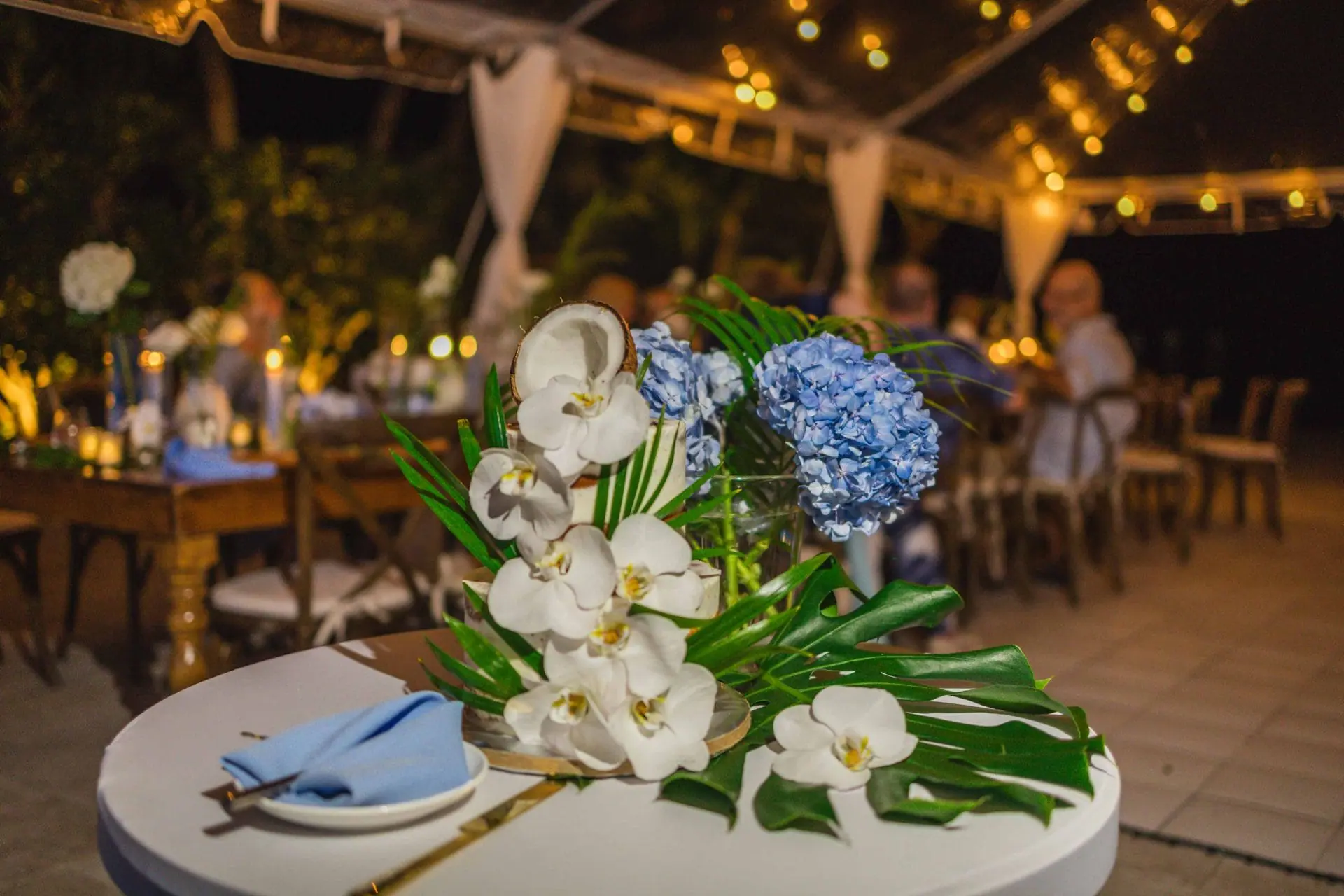 A table centerpiece with white orchids and blue hydrangeas, under a canopy with string lights at night.