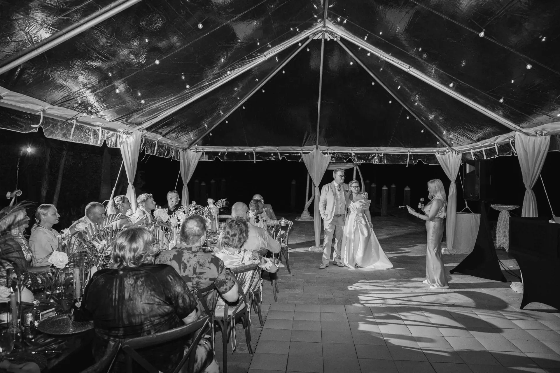 A bride and groom stand under a transparent tent while a woman makes a speech to seated guests at a wedding reception.