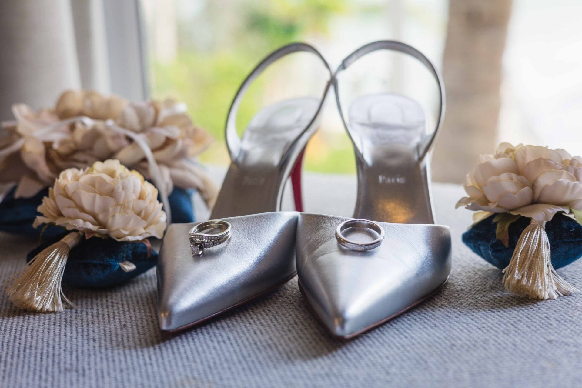 Silver high heels displayed with wedding rings and cream floral boutonnieres on a gray surface.